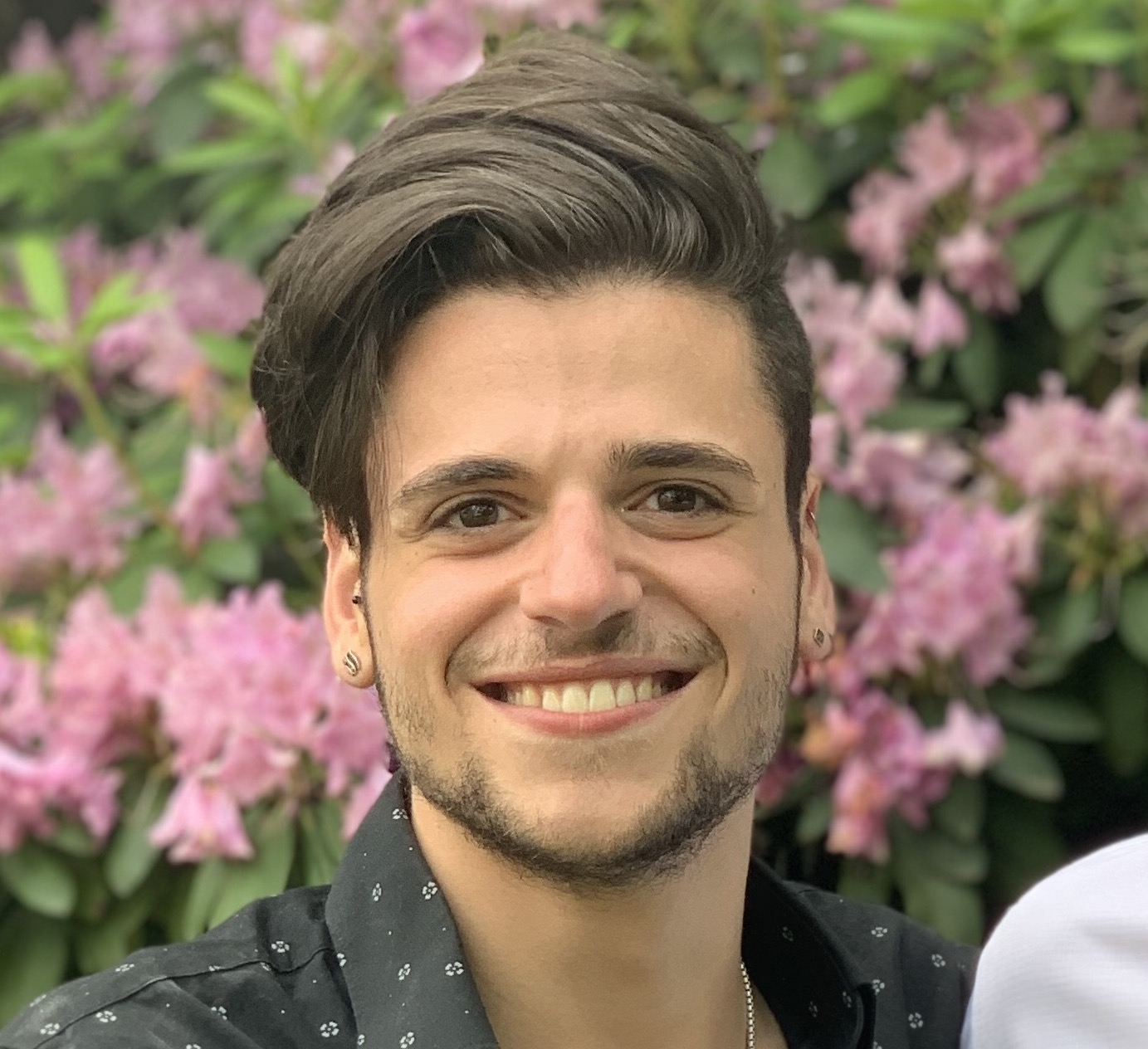 Joseph Guzzardo, a man with thick medium length brown hair swooped to the side, brown eyes, and fair skin, wearing a block button down smiling in front of a bush with pink flowers.